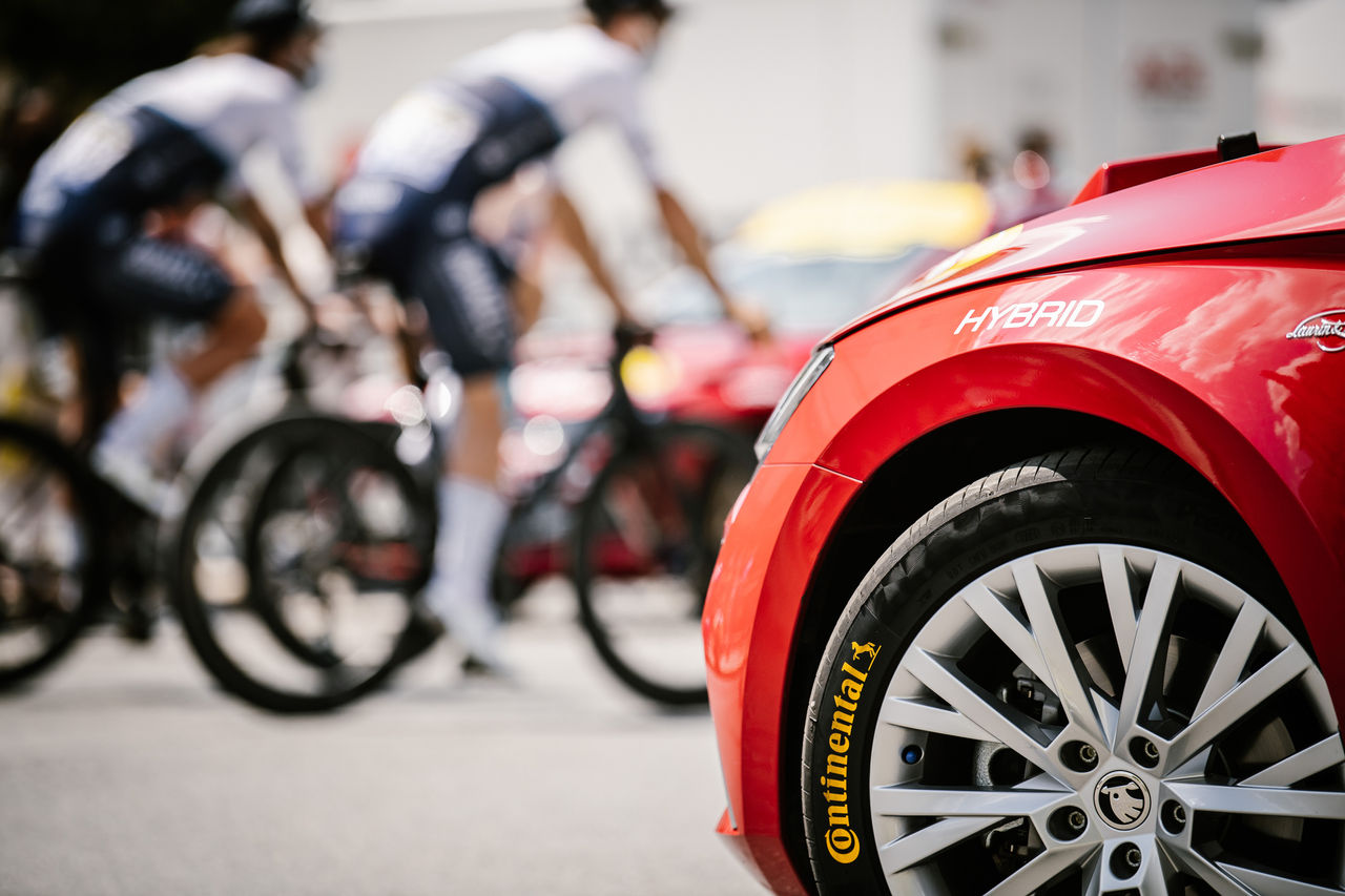 Foto de un carro rojo con llantas Continental durante el evento del Tour de Francia, con personas en bicicleta al fondo.