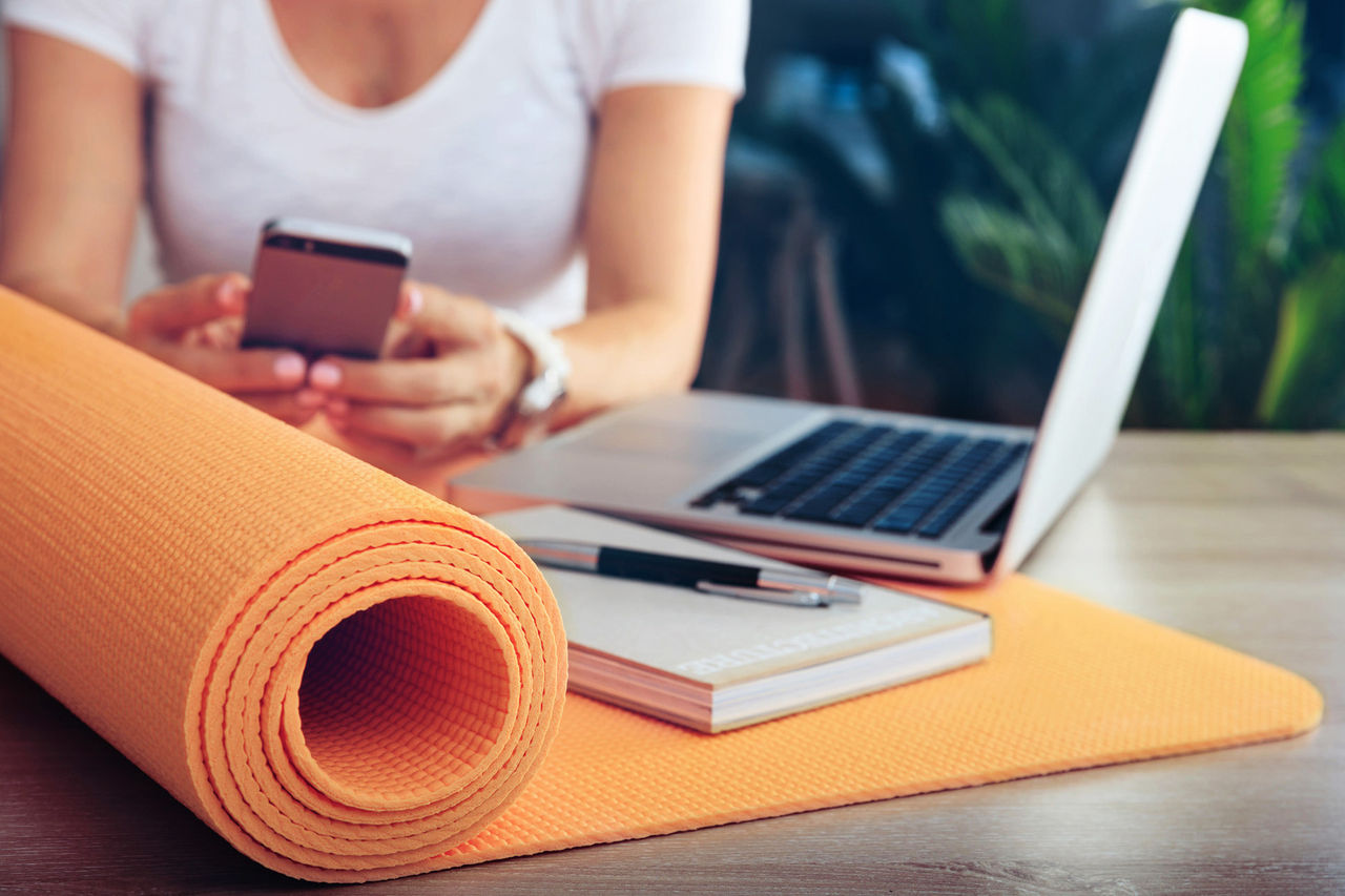 A half-rolled orange yoga mat on a table with a book, a pen, and a laptop, with a woman holding a cell phone in the background