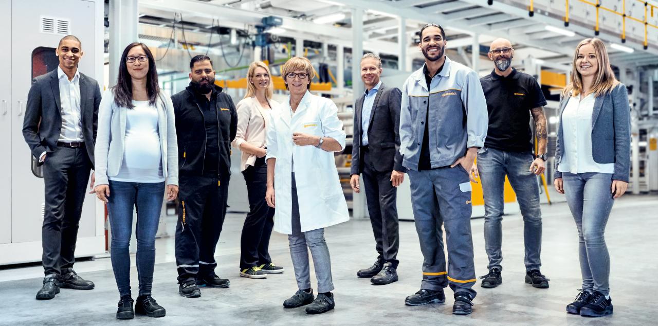 Two men in suits, two men in work clothes, a woman in a suit, a woman in a lab coat, a woman and a man in casual work clothes, and a pregnant woman standing in a production facility smiling