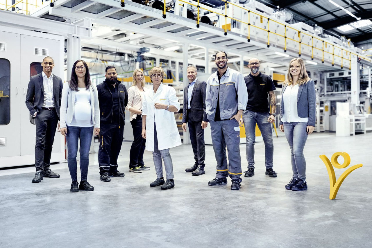 A diverse group of nine people, including men and women in business and work attire, stand smiling in a spacious, modern industrial facility with machinery in the background.