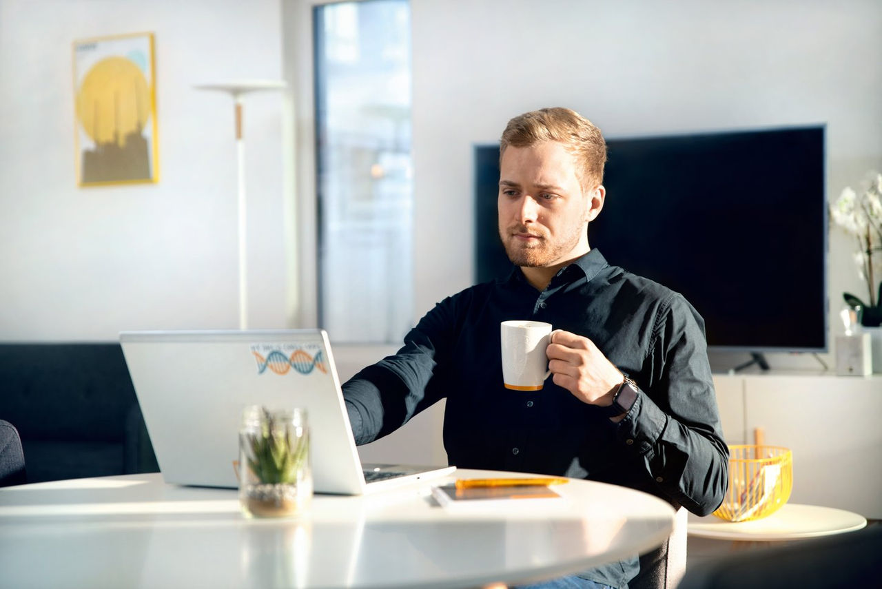 A man in a black shirt sits at a white table using a laptop, holding a white mug. Sunlight fills the modern room, with a TV, plant, and wall art visible in the background.