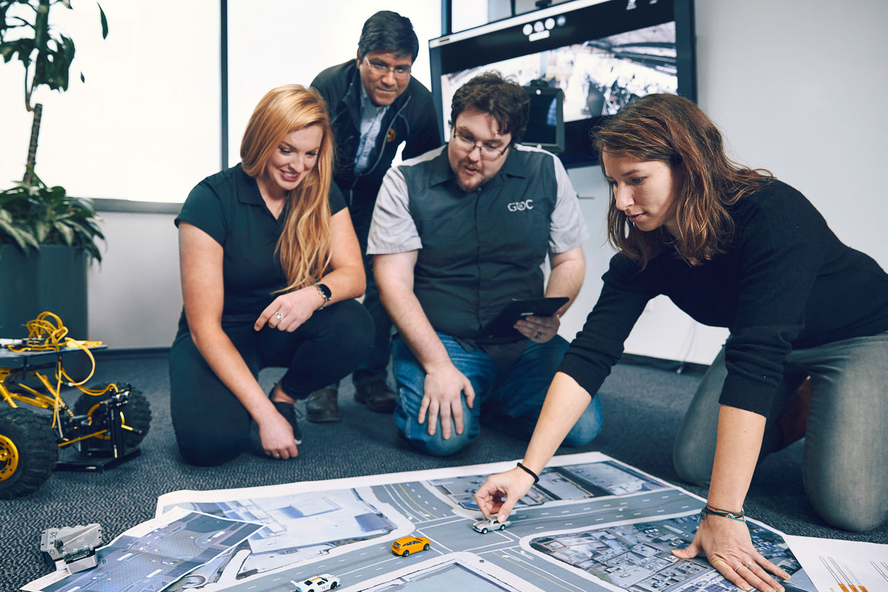 Four people kneel around a map of roads on the floor, using toy vehicles to simulate traffic. A model construction vehicle and plants are nearby, suggesting a collaborative planning or engineering session.