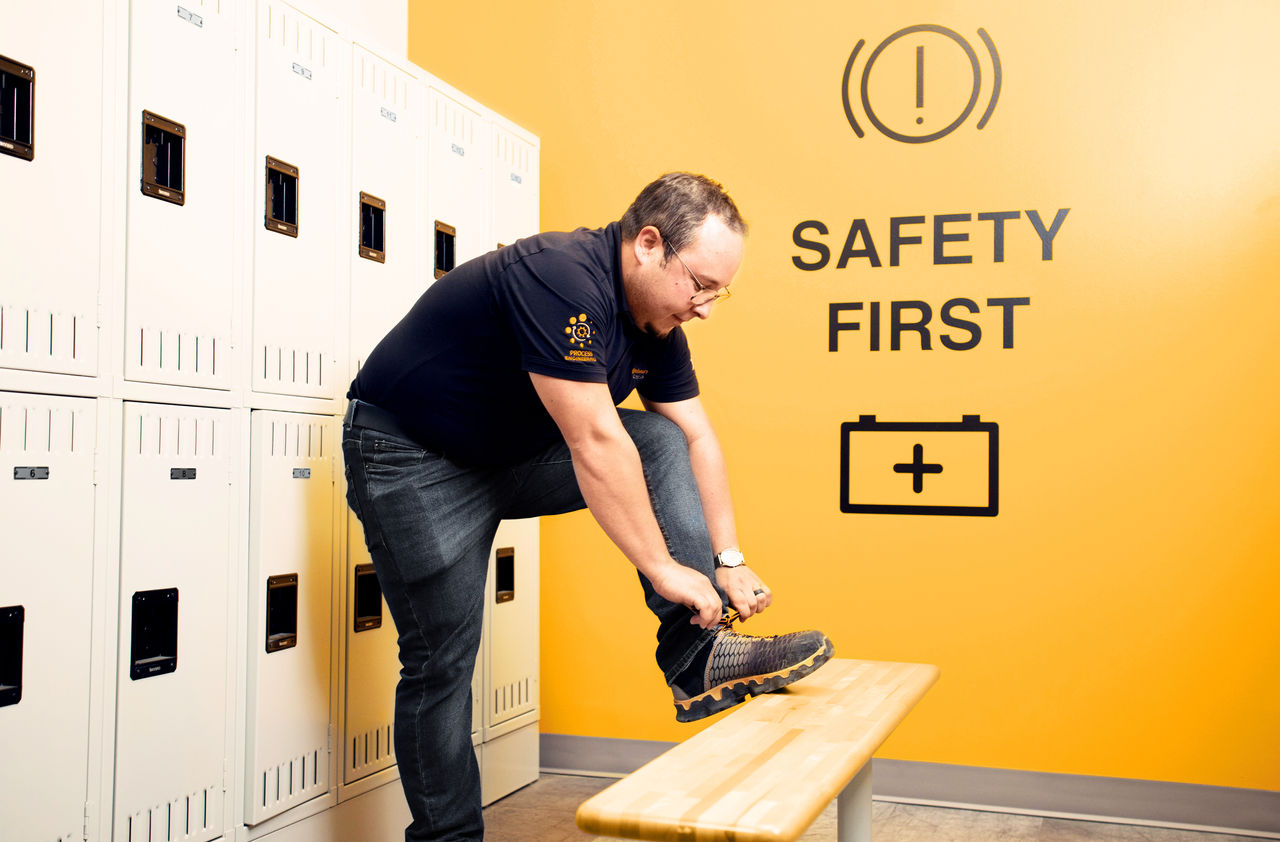 A man in a black shirt puts on safety boots in a locker room with beige lockers. The yellow wall behind him displays a "SAFETY FIRST" sign with warning and medical icons.