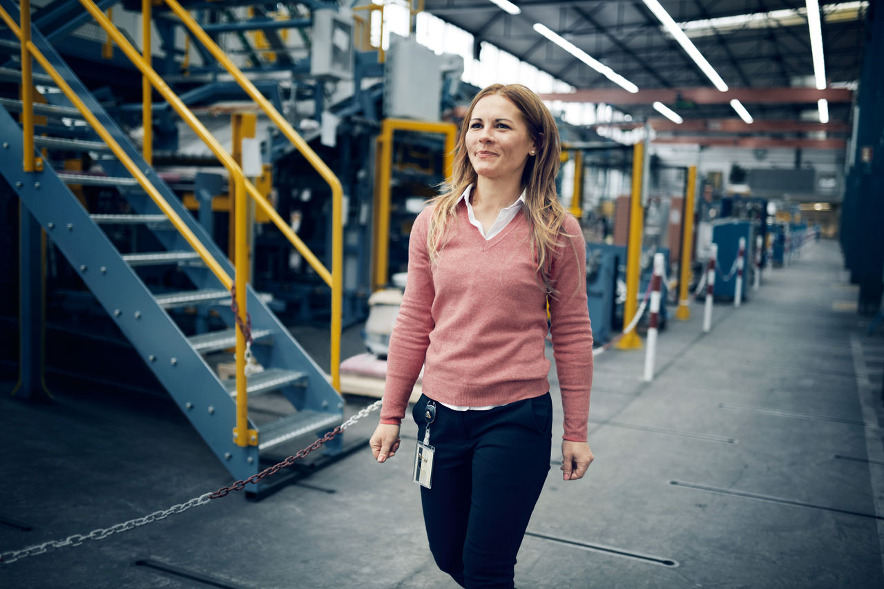 A woman wearing a pink sweater and dark pants walks confidently in a large industrial factory, with machinery and metal staircases in the background.
