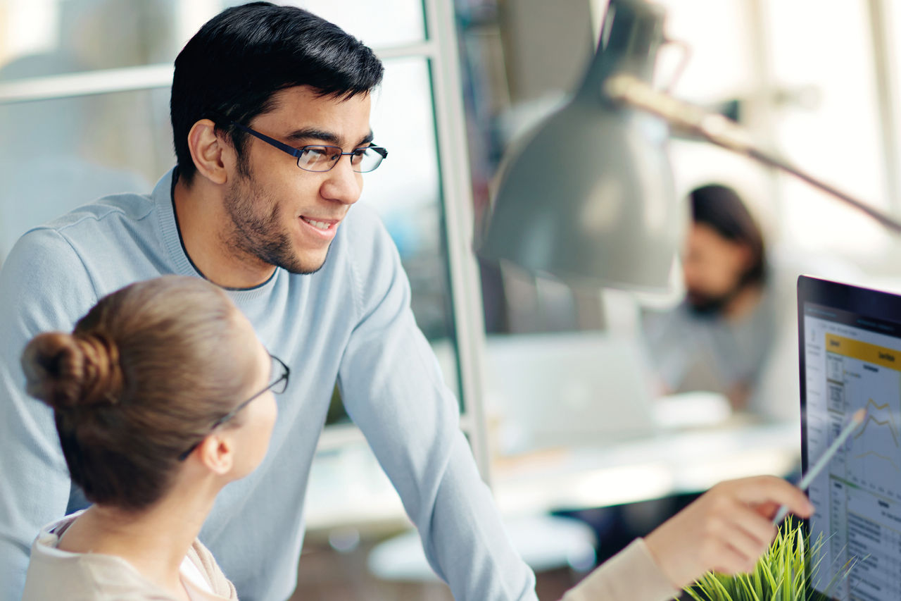 A man with glasses is standing at a desk looking at what a woman with glasses is showing him sitting on a display