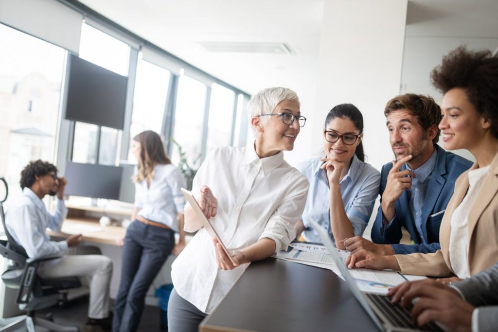 A group of professionals gathers around a desk in a modern office, discussing work and looking at documents and a laptop, while two colleagues converse in the background.