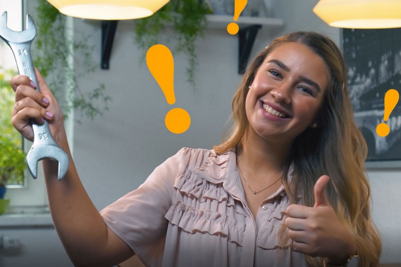 A smiling woman holds up a large wrench in one hand.