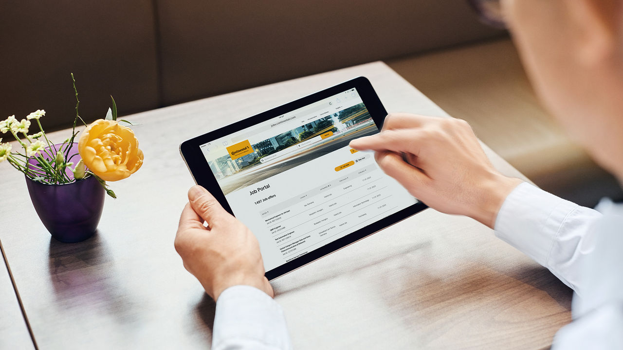 A person using a tablet to browse job postings at a table, with a yellow flower in a purple vase nearby. The screen displays a job portal website with listings and a yellow “Apply now” button.