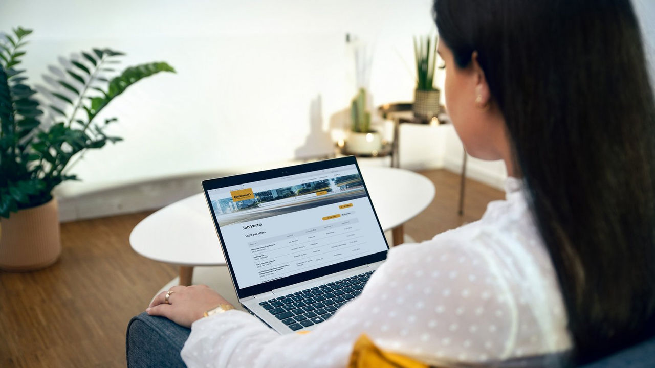 A woman sits in a modern, well-lit room looking at a laptop screen displaying a job portal website with listings. A round white table, plants, and decor are visible in the background.