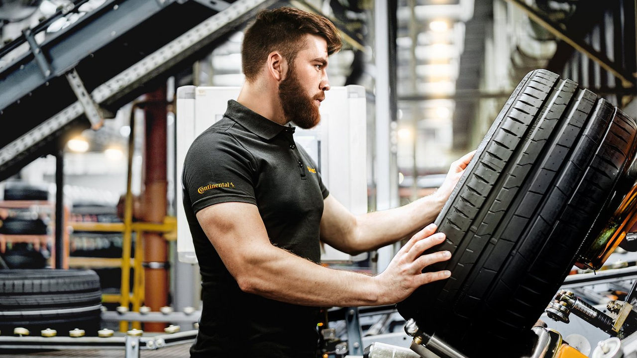 A man wearing a black Continental shirt inspects a large car tire in a factory setting, surrounded by industrial equipment and tires.