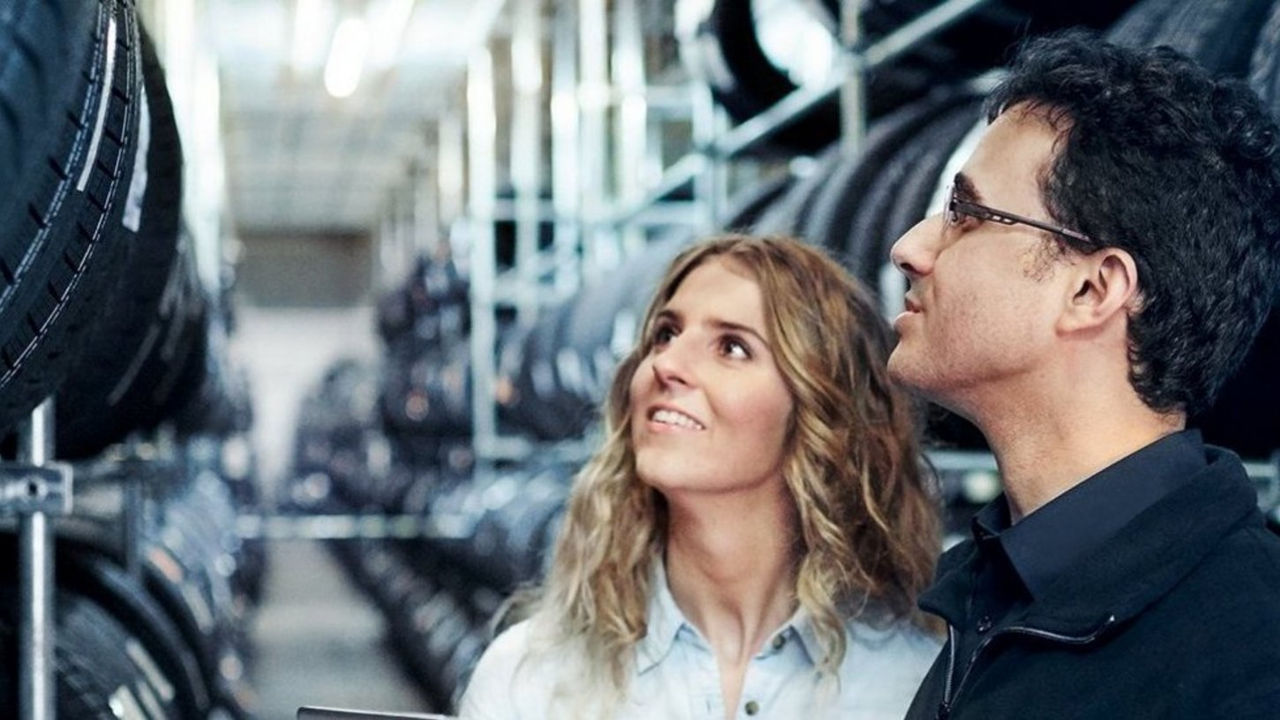 A woman and a man stand in a warehouse lined with shelves of tires, looking up and smiling, possibly inspecting or discussing the inventory.
