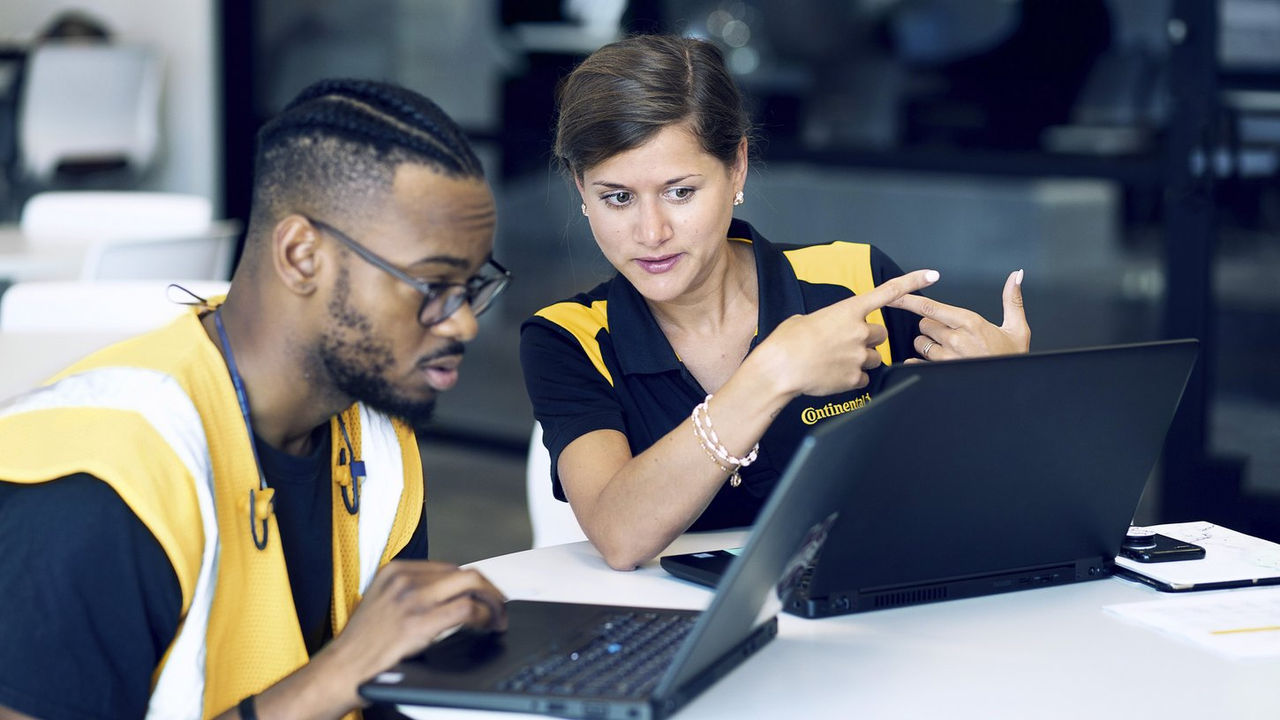 Two coworkers sit at a table with laptops. The woman, wearing a black and yellow shirt, gestures while talking to the man, who wears glasses and a yellow vest. They appear focused and engaged in discussion.