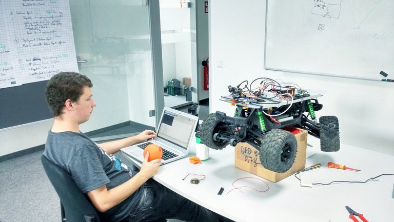 A person sits at a desk working on a laptop, holding an orange object. Next to them is a large, partially assembled remote-control car with exposed wires, placed on a box. Tools and electronic parts are scattered on the table.