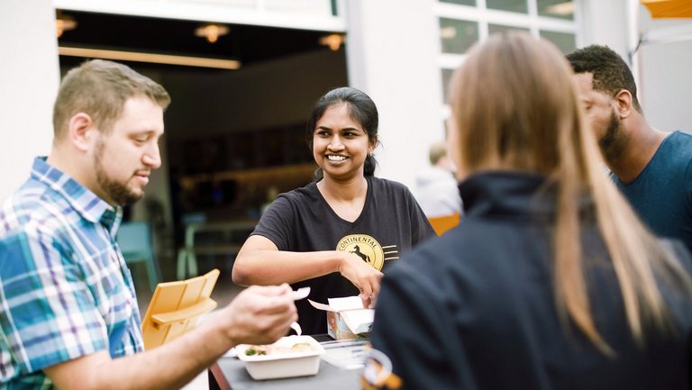 A group of four people gather around a tall table outdoors, eating and talking. One woman in a black shirt smiles brightly, while others engage in conversation with food containers in front of them.