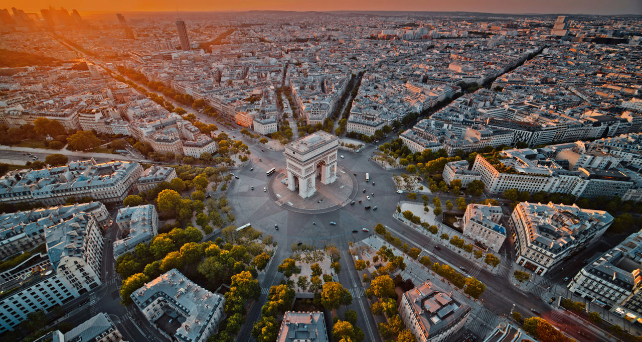 Aerial view of the Arc de Triomphe at the center of Paris, where wide avenues spread outward in all directions, glowing in warm sunset light