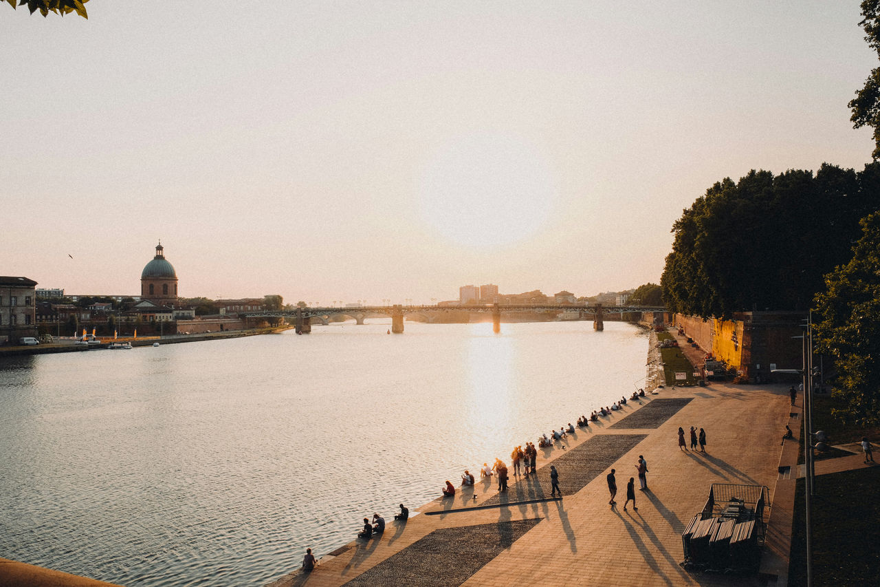 People stroll and relax along the Garonne River in Toulouse at sunset, with the dome of La Grave hospital visible across the water. 
