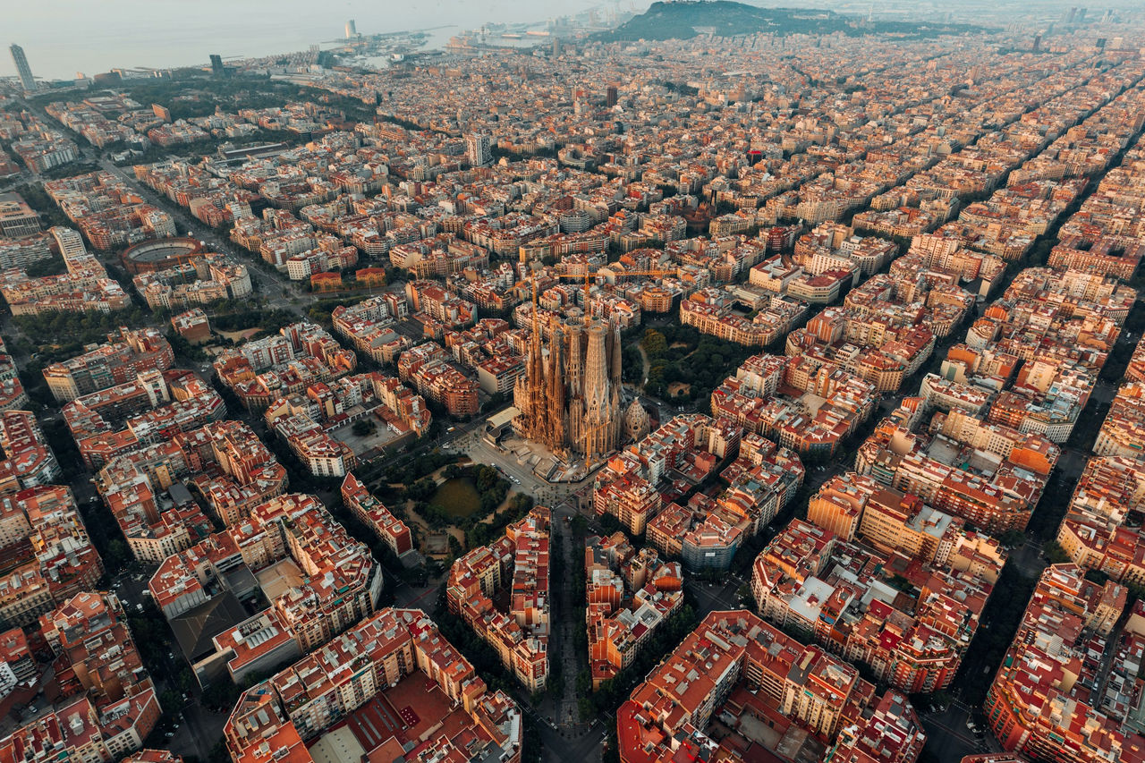 Aerial view of Sagrada Família rising above Barcelona, with warm golden light from sunrise casting long shadows across the city’s grid and highlighting the surrounding rooftops.