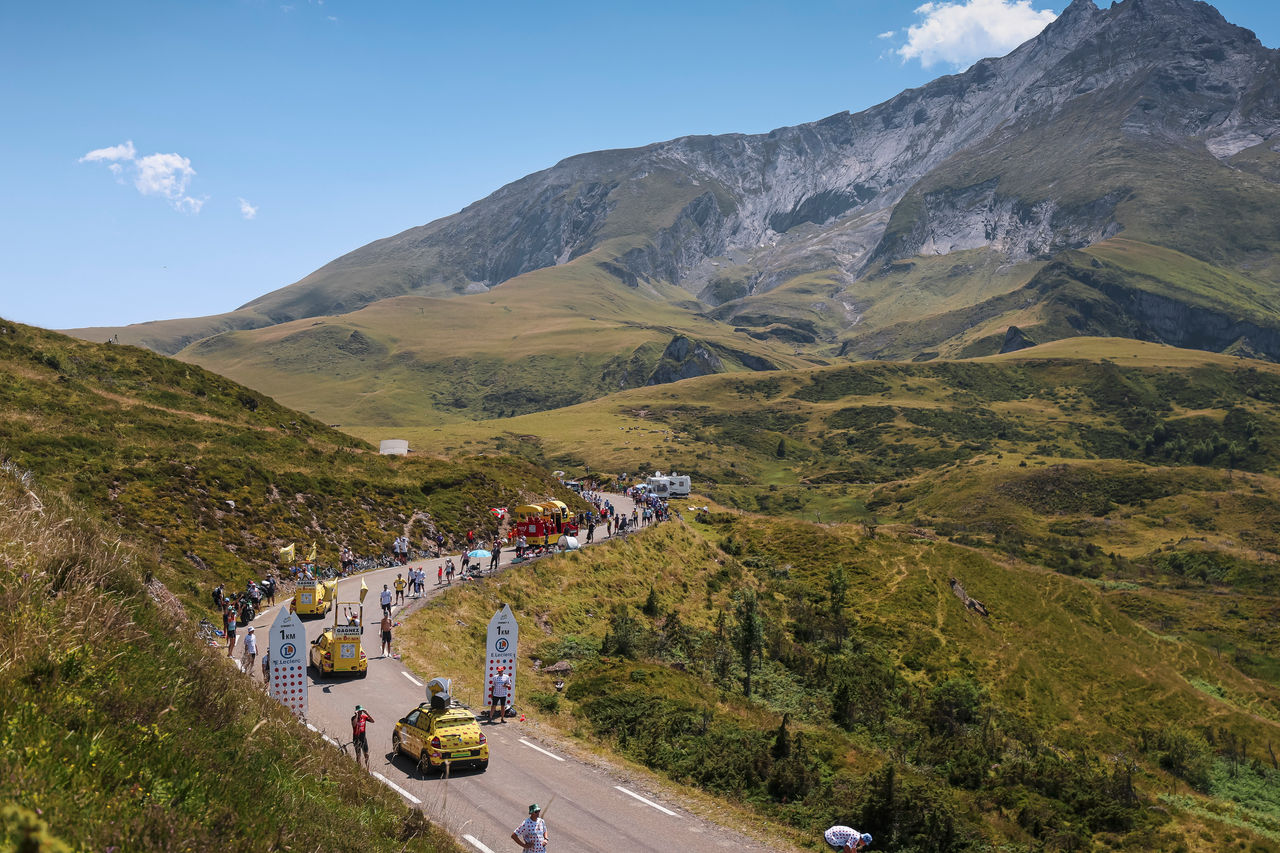 A winding mountain road lined with spectators and support vehicles, with rolling green hills and a towering rocky peak under a clear blue sky.