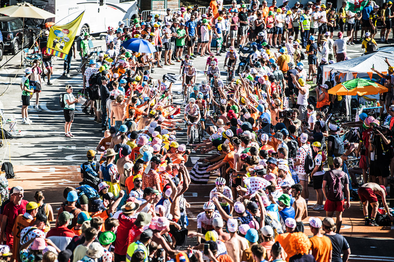 A large crowd lines a narrow road, cheering as cyclists ride past during a race.