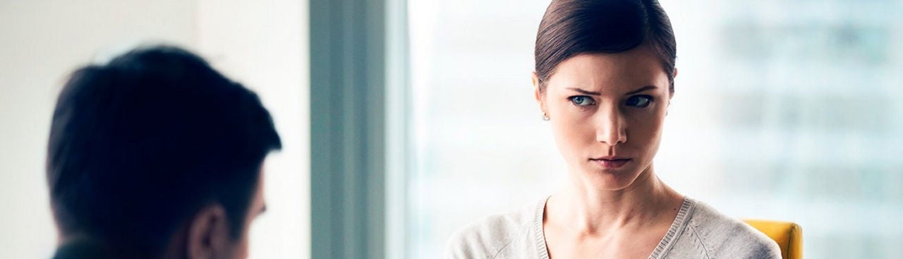 A woman with brown hair pulled back is sitting indoors, looking sideways with a serious expression at a man whose back is to the camera.