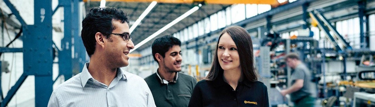 Three people, two men and one woman, walk and talk together inside a bright, industrial factory or warehouse, with machinery and equipment visible in the background.