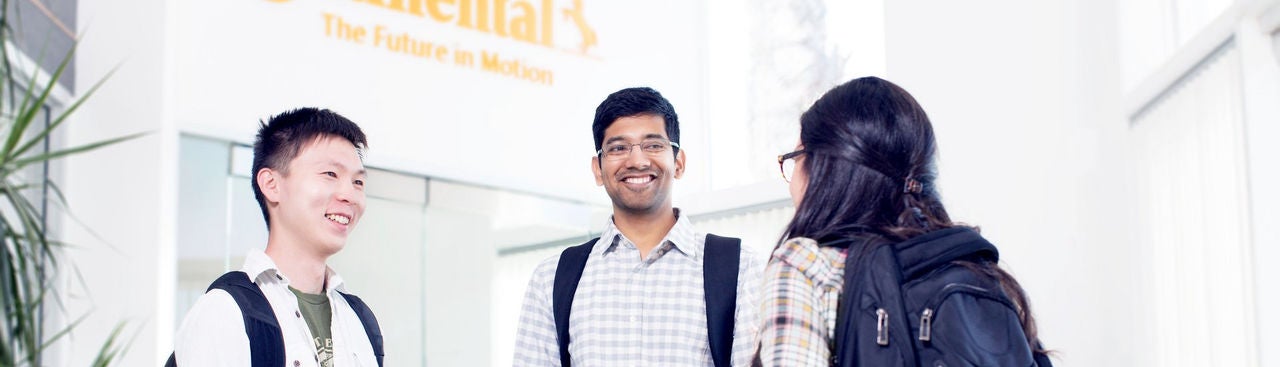 Two young men and a young woman wearing casual business attire standing in a lobby and talking to each other in front of an elevator 
