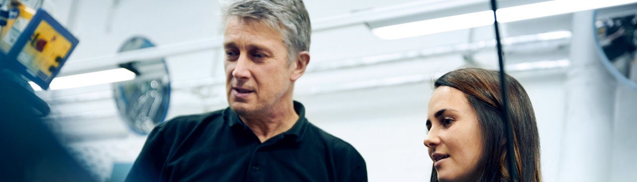 A man and a woman, both focused, look at something off-camera in a bright, modern workspace with industrial elements in the background.