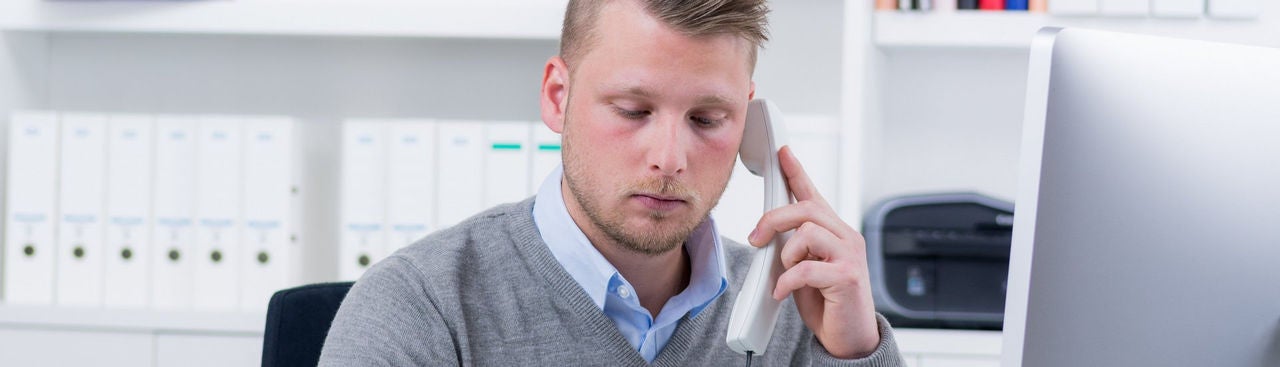 A man in a gray sweater sits at a desk in an office, talking on a corded phone while looking at a computer monitor. 