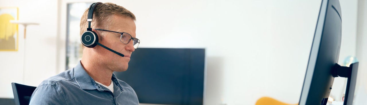 A man wearing glasses and a headset sits at a desk, looking at a computer monitor, appearing focused. 