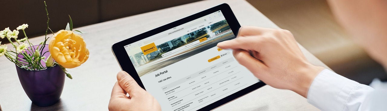 A person uses a tablet displaying a job portal website at a desk, with a small vase containing a yellow flower and greenery placed nearby.