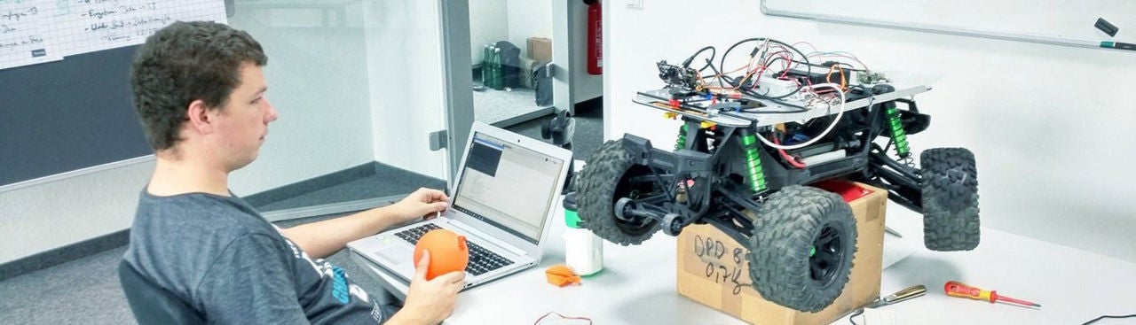 A person sits at a desk working on a laptop, holding an orange object. Next to them is a large, partially assembled remote-control car with exposed wires, placed on a box. Tools and electronic parts are scattered on the table.