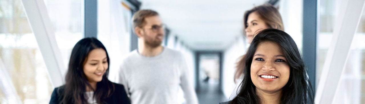 Four young professionals walk together in a bright, modern hallway. The focus is on a smiling woman in the foreground, while three other people walk and talk behind her.