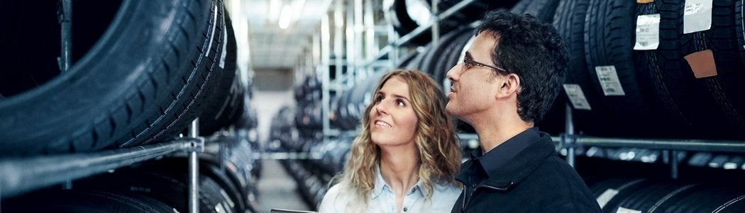 Two people, a woman and a man, are standing in a warehouse aisle lined with car tires on shelves. They are looking up and appear to be inspecting or selecting tires. The setting is brightly lit and industrial.