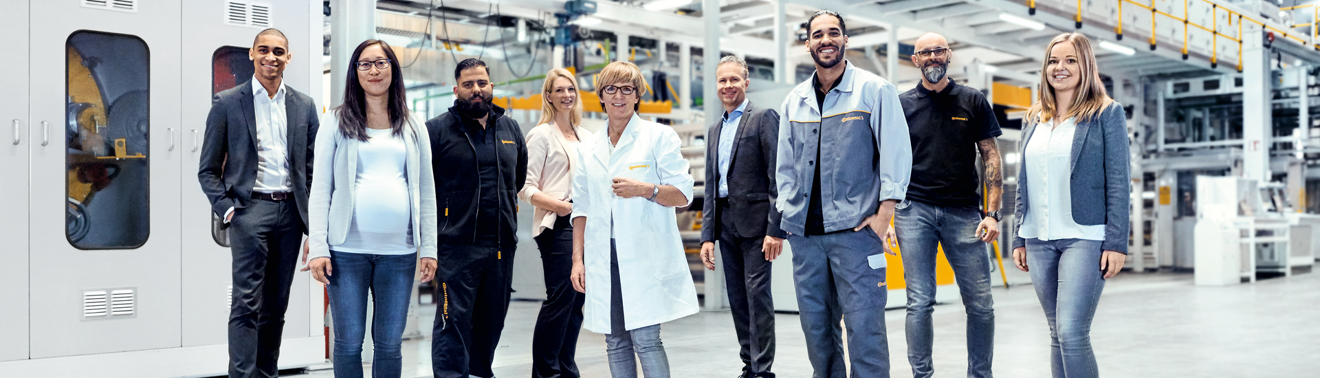 Two men in suits, two men in work clothes, a woman in a suit, a woman in a lab coat, a woman and a man in casual work clothes, and a pregnant woman standing in a production facility smiling 