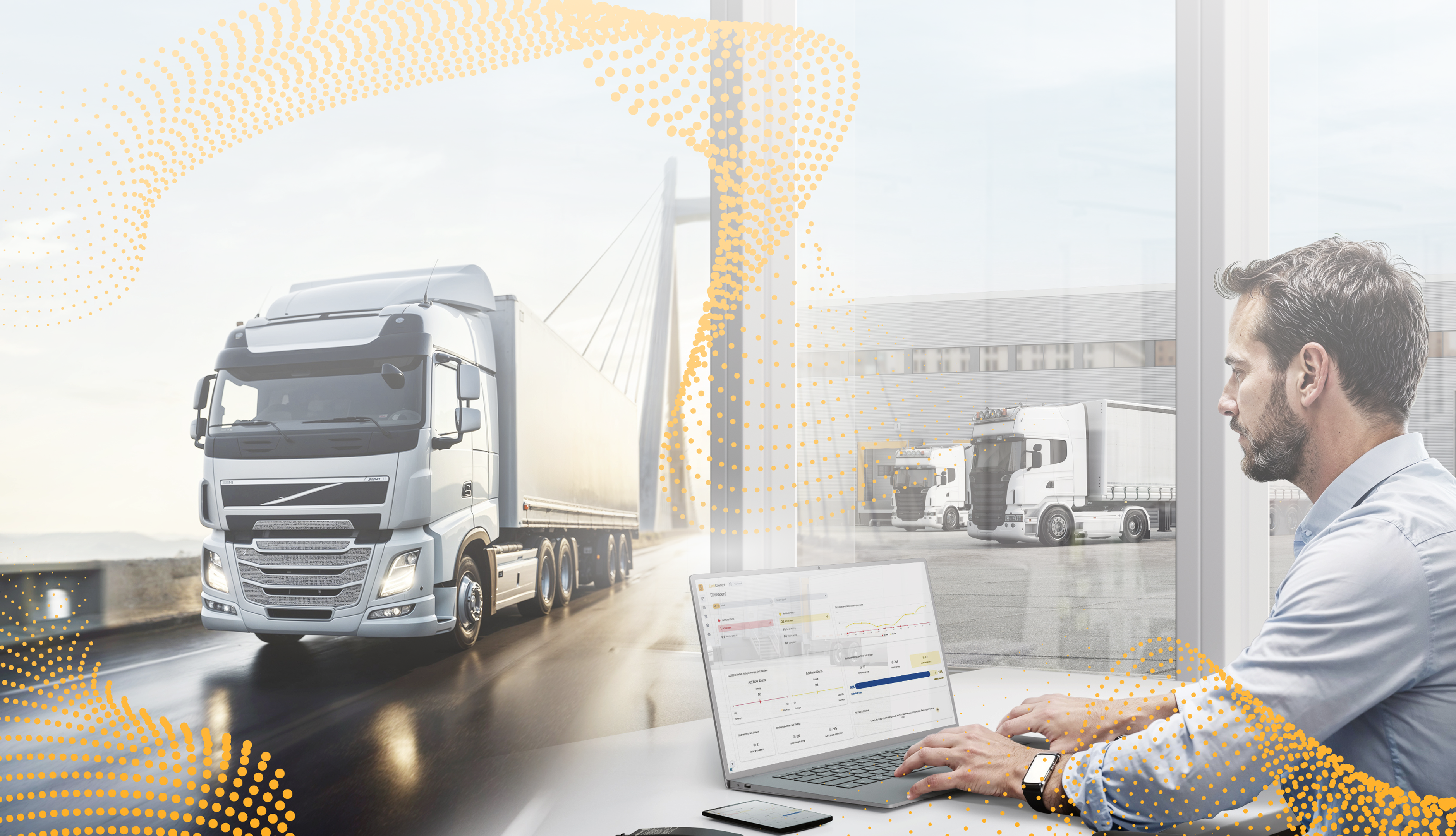 A man works on a laptop near a large window, overlooking a parking area with trucks. A semi-truck drives on a bridge outside.
