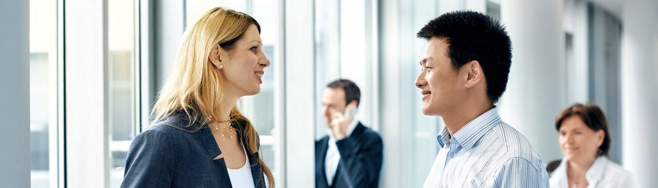 Two professionals, a woman and a man, are having a conversation by large windows in an office. In the background, a man is on the phone and a woman is walking. The atmosphere appears bright and businesslike.