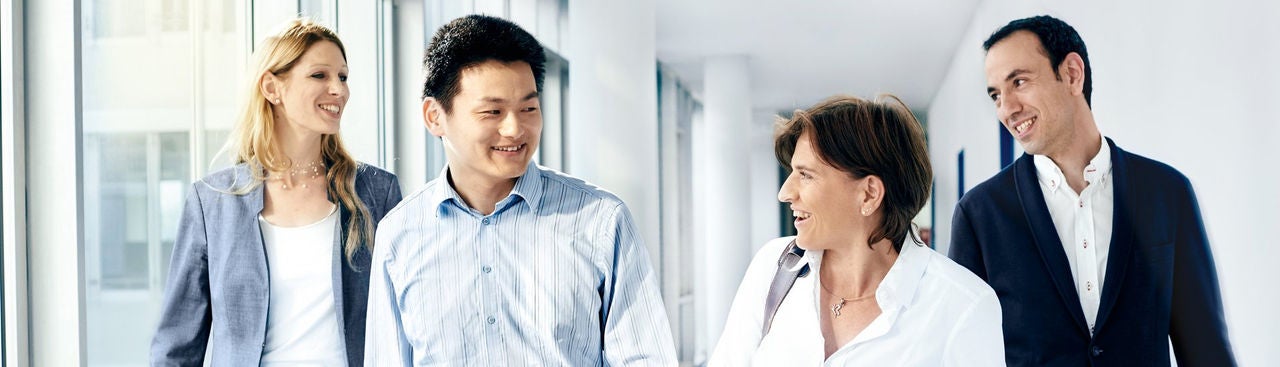 Four professionally dressed people are walking down a bright hallway, smiling and talking together. Two women and two men appear to be enjoying a friendly conversation in a modern office setting.