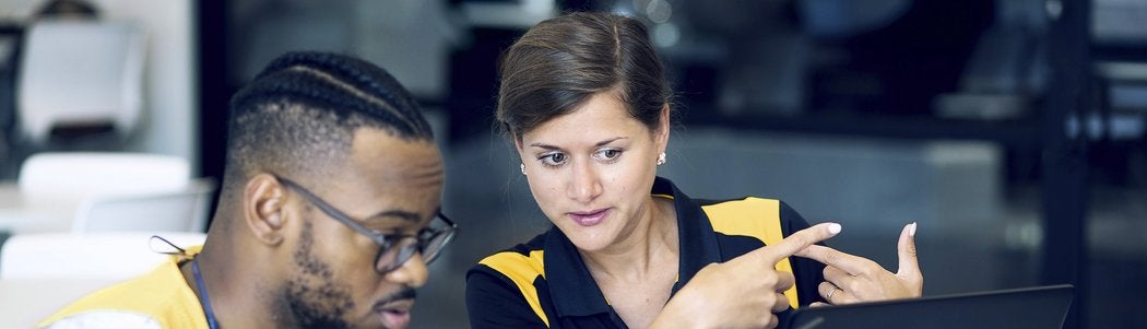 A woman points at a laptop screen while talking to a man beside her. Both are seated indoors and appear to be working or studying together, focused on the device in front of them.