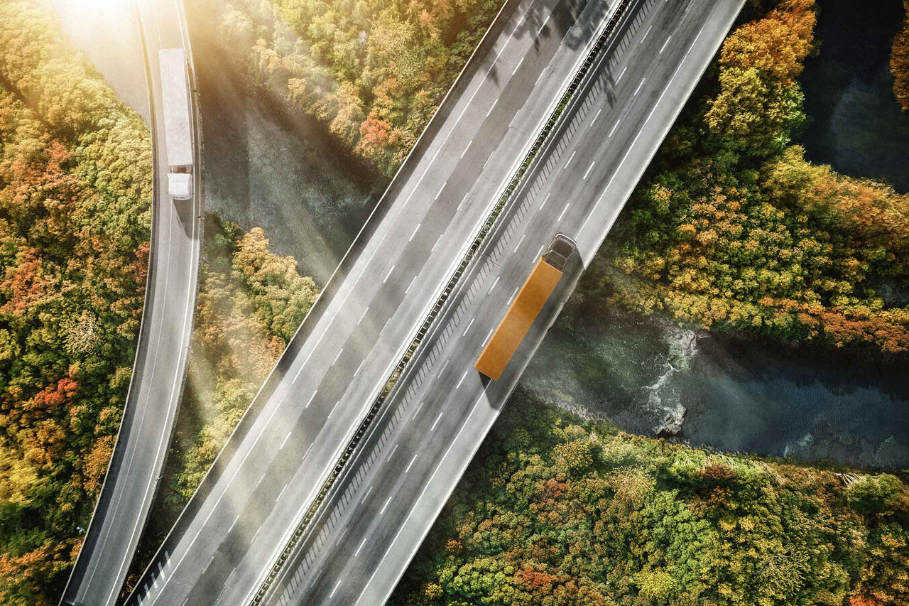 Servicios y soluciones digitales para camiones y ómnibus (Muestra una vista aérea de una autopista que corre sobre un río sinuoso y rodeada de vegetación. Hay dos carriles de tráfico visibles; uno de ellos, a la derecha, está ocupado por un camión largo, predominantemente gris, con un balde naranja. La carretera es de asfalto ligero y los carriles están marcados con líneas blancas. A su alrededor, el paisaje es rico en tonos de verde, naranja y amarillo, lo que sugiere que es otoño. El río de abajo es de un tono más oscuro y serpentea suavemente entre la vegetación, mientras que la luz del sol crea un brillo en las partes superiores de la imagen, lo que indica que es un día claro.)