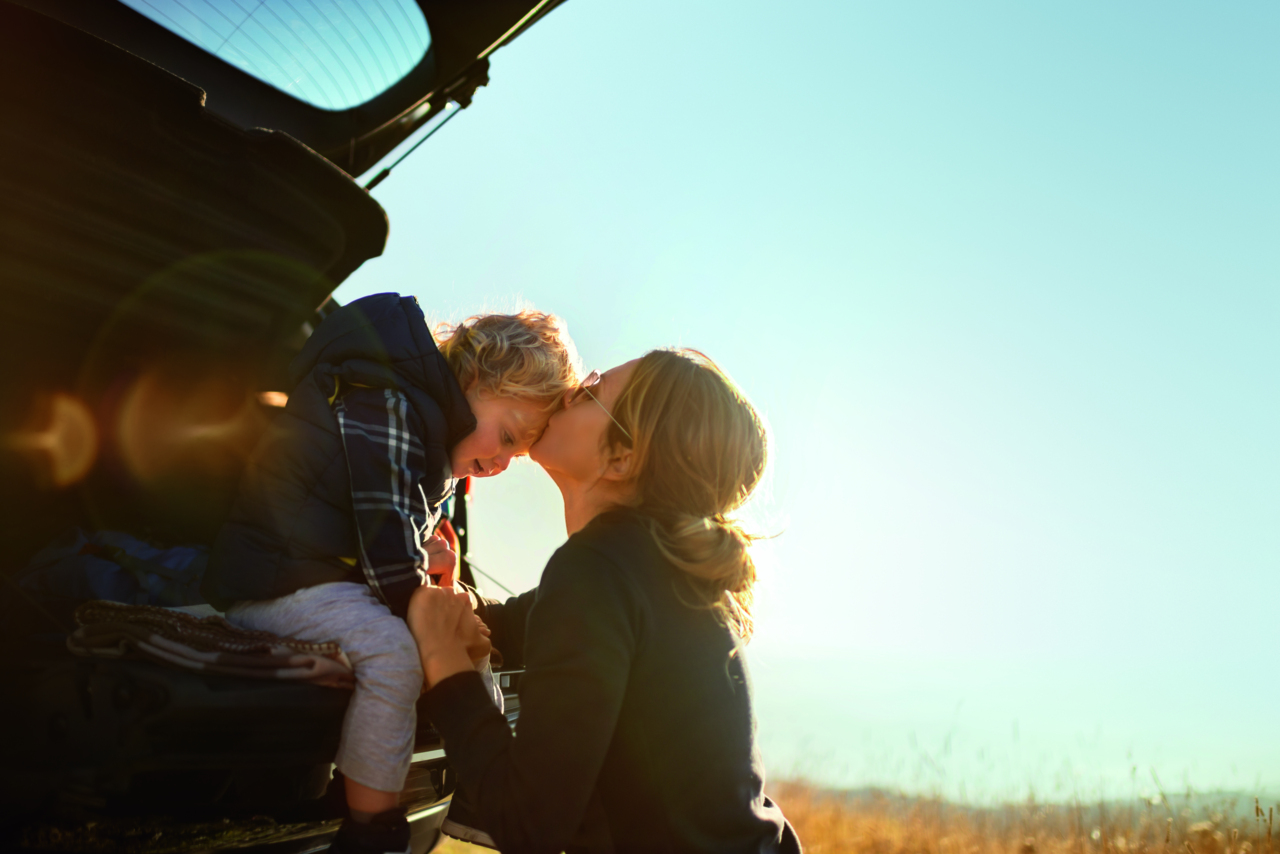 Mother and little son travel by car in mountains.Little boy sitting in car trunk while they makes a break and mother gives him a kiss.