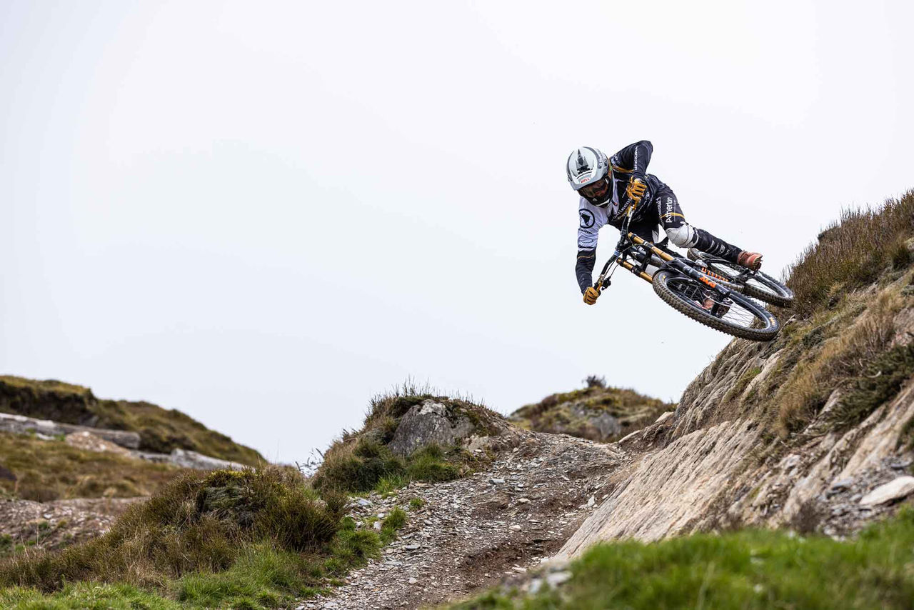 Llanta Continental para bicicleta (La imagen captura a un ciclista realizando una maniobra en un sendero de mountain bike. El ciclista está inclinado hacia un lado, con el cuerpo en un ángulo acentuado, destacándose sobre un terreno rocoso y accidentado. Lleva casco y ropa de protección, y la bicicleta con llantas Continental parece estar a alta velocidad, indicando un ambiente de adrenalina y aventura. Al fondo, se mezcla rocas y vegetación, con un cielo claro que proporciona un contraste al escenario. El ciclista parece estar en movimiento rápido, resaltando la habilidad y la técnica necesarias para navegar por este terreno desafiante.)