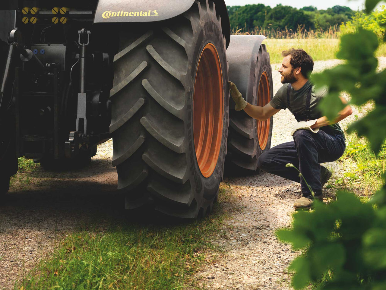 Llanta Continental Agrícola (La imagen muestra a un hombre agachado junto a un tractor, inspeccionando una de las llantas, que son de tipo agrícola Continental. Él lleva una camiseta oscura y guantes, y tiene cabello corto y barba. La rueda del tractor es grande, con un patrón de ranuras profundas en la llanta, mientras que el borde de la rueda es de color naranja. El escenario es al aire libre, con un camino de tierra y hierba alrededor; al fondo, hay vegetación verde que sugiere un ambiente rural. La luz es natural, indicando un día soleado. La atmósfera transmite una sensación de trabajo y conexión con la naturaleza.)