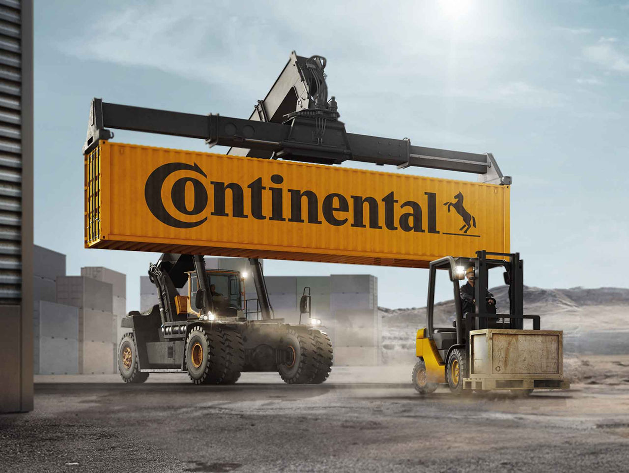 Llantas Continental Industriales, Portuarias, Aeroportuarias, Superelásticas y Sólidas (La imagen muestra un escenario industrial donde dos vehículos de carga con llantas Continental están en acción. En el centro, una autoelevadora grúa grande que levanta un contenedor metálico amarillo con el logotipo de "Continental" en el costado. La grúa tiene un brazo largo y robusto, demostrando su capacidad para levantar cargas pesadas, mientras que el contenedor permanece suspendido en el aire. A la derecha, una autoelevadora amarilla está en movimiento, transportando material en una plataforma inferior, que parece ser de madera o metal. El fondo está compuesto de estructuras de concreto, con un cielo claro y algunas nubes suaves. El ambiente transmite una sensación de actividad intensa y eficiencia operativa. El suelo muestra marcas y polvo, indicando el tráfico de vehículos pesados.)