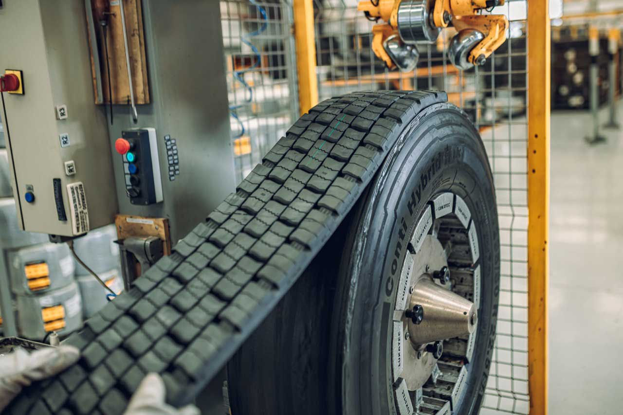 (Máquina automatizada aplicando una nueva banda de rodadura en una llanta de carga de Continental, en un entorno dentro de una fábrica. Se ven manos usando guantes)