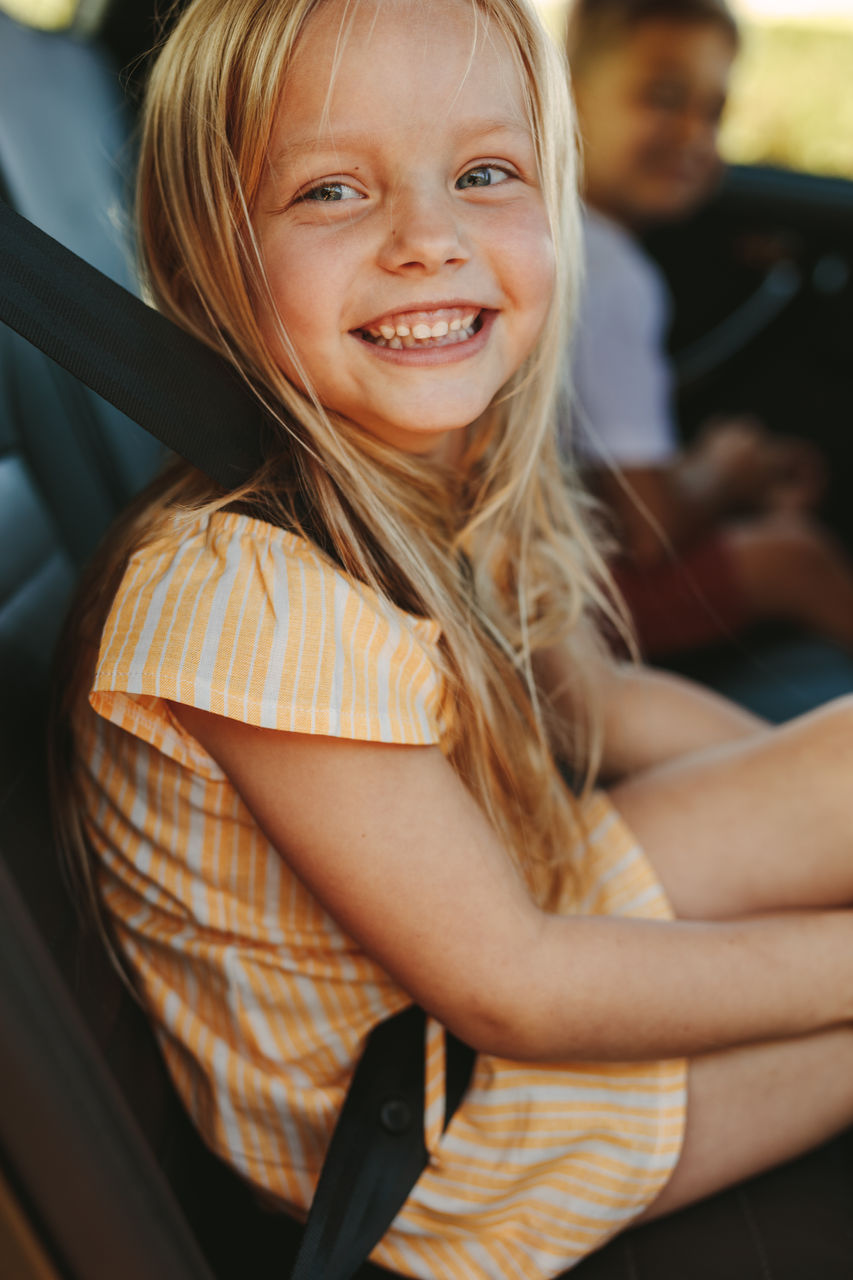 Close-up of a cute girl sitting in backseat of car wearing seatbelt. Cheerful girl enjoying travelling by a car.