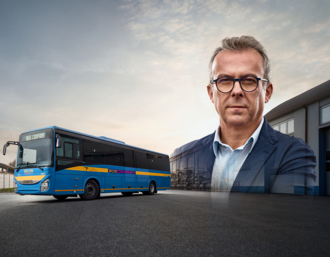 A confident middle-aged man in glasses stands in front of a garage, with a blue and yellow bus labeled "BUS COMPANY" parked nearby under a partly cloudy sky.