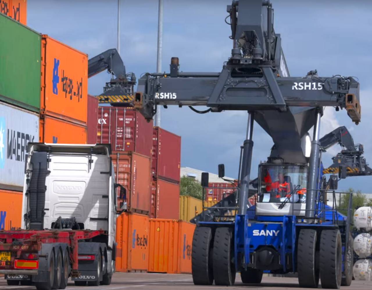 A large reach stacker lifts a shipping container in a busy container yard, with colorful stacked containers and a white Volvo truck on the left, under a partly cloudy sky.