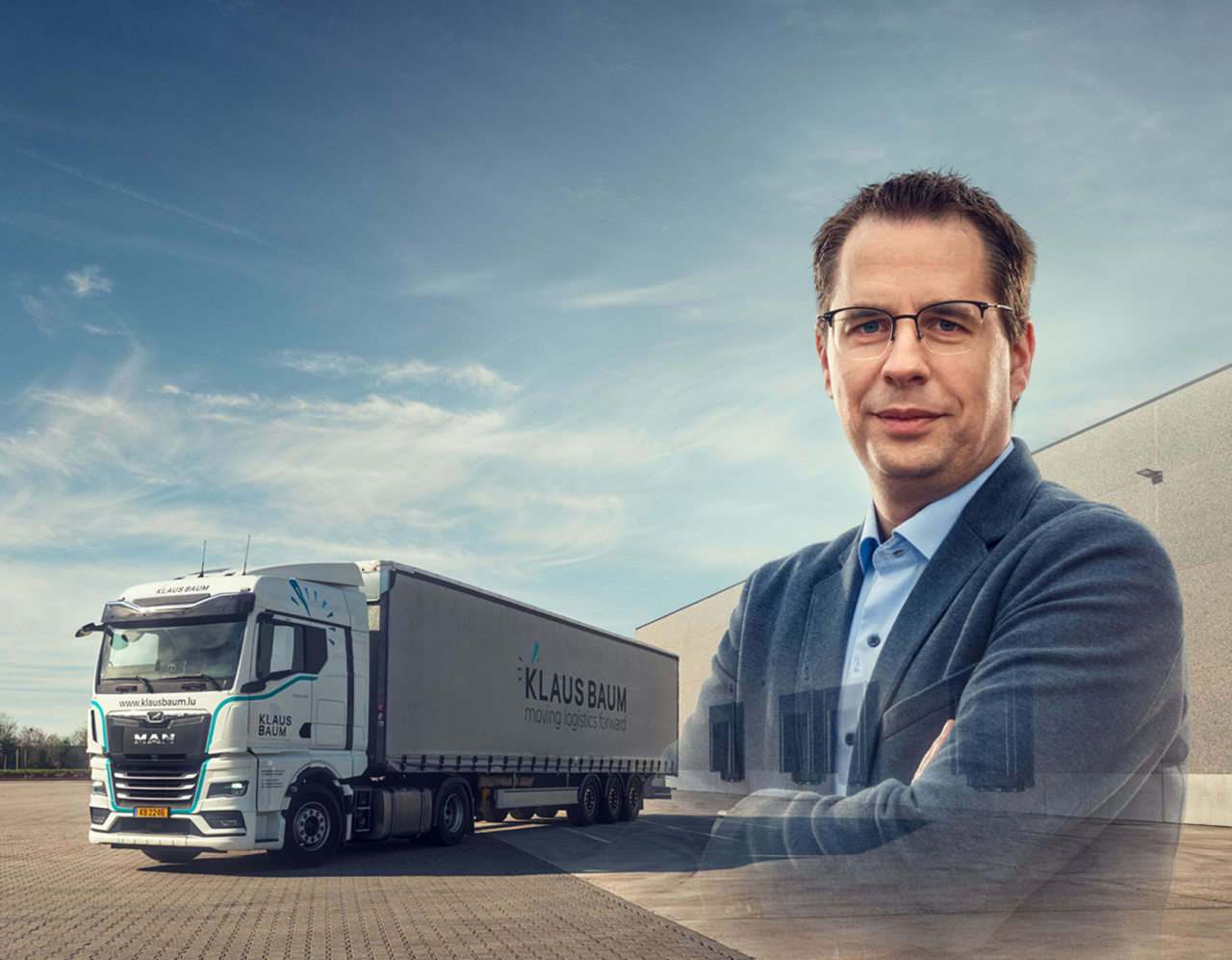 A man in glasses and a suit jacket stands with arms crossed in front of a large Klaus Baum logistics truck parked outside a warehouse under a blue sky.