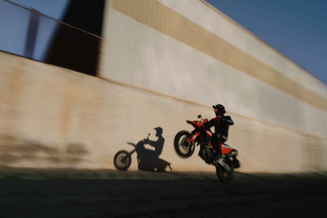 Llanta Continental para motocicleta (La imagen muestra a un motociclista realizando una maniobra de "wheelie", en la que la moto se mantiene en pie solo sobre la rueda trasera. El piloto, con casco y ropa oscura, parece desafiar la gravedad mientras la moto, de color naranja y con llantas Continental, se eleva del suelo. El fondo es una pared de color clara y texturizada, creando un contraste impresionante. La sombra del motociclista se proyecta en la pared, destacando el movimiento rápido y audaz. La atmósfera transmite una sensación de velocidad y adrenalina, con un cielo azul al fondo, sugiriendo un día claro y soleado.)