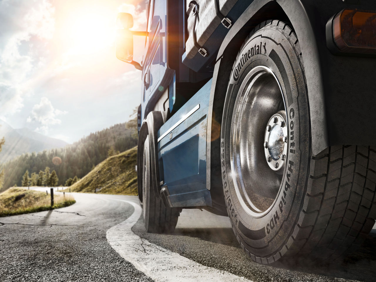 Llanta Continental para Camión y Bus (La imagen muestra un camión grande en una carretera sinuosa rodeada de un paisaje montañoso. La perspectiva es desde el lado del camión en la parte trasera, destacando una de sus llantas traseras. La llanta, de la marca Continental, es robusta y tiene un diseño profundo, indicando que es adecuada para carreteras, modelo Conti Hybrid HD3. El camión en sí está pintado de azul y presenta detalles plateados, reflejando la luz del sol que viene desde un ángulo de la imagen. El cielo está parcialmente nublado, con rayos de sol penetrando las nubes, creando un ambiente brillante y natural. Al fondo, se pueden ver colinas verdes, agregando un toque de tranquilidad a la escena, mientras la carretera se curva hacia la imagen